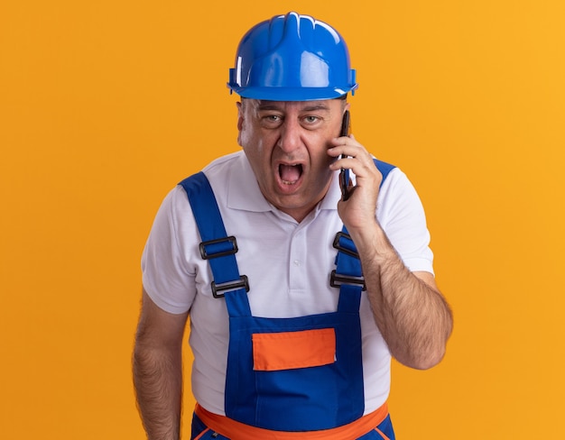Boze volwassen bouwersman in uniform schreeuwt tegen iemand op de telefoon die op oranje muur wordt geïsoleerd