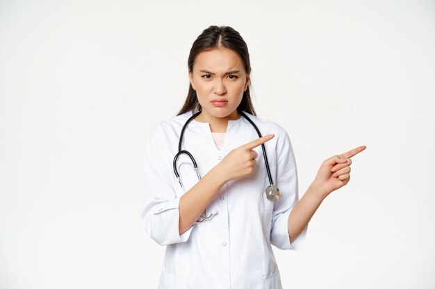 Boze vrouwelijke arts, aziatische arts in medisch gewaad en stethoscoop, wijzende vingers naar rechts en fronsend woedend, starend teleurgesteld, witte achtergrond
