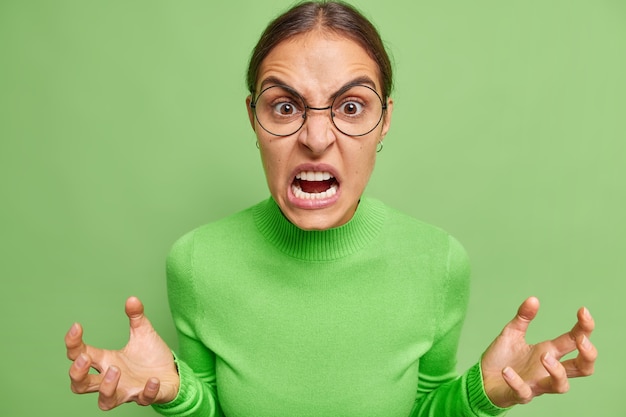 Gratis foto boze woedende vrouw kijkt met woede gebaren en fronst gezicht geïrriteerd met iets draagt ronde bril casual coltrui poses tegen felgroene muur