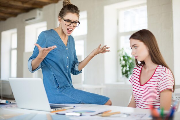 Boze zakenvrouw in brillen leunend op bureau terwijl ze schreeuwen op werknemer in modern kantoor