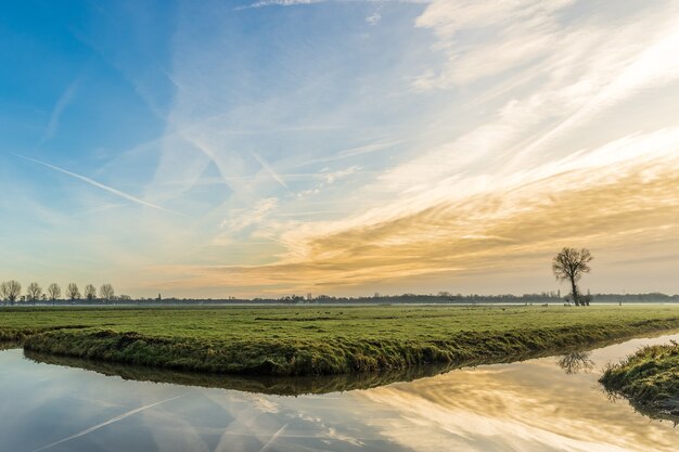 Brede opname van een met gras begroeid veld met een watermassa als gevolg van de prachtige zonsondergang en de lucht