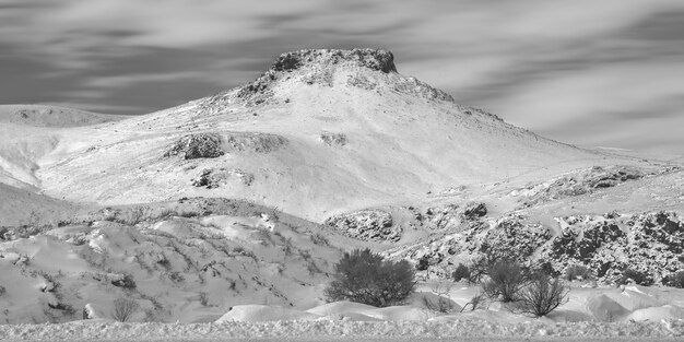 Breed grijswaarden shot van besneeuwde heuvels en een berg in de verte met een bewolkte hemel