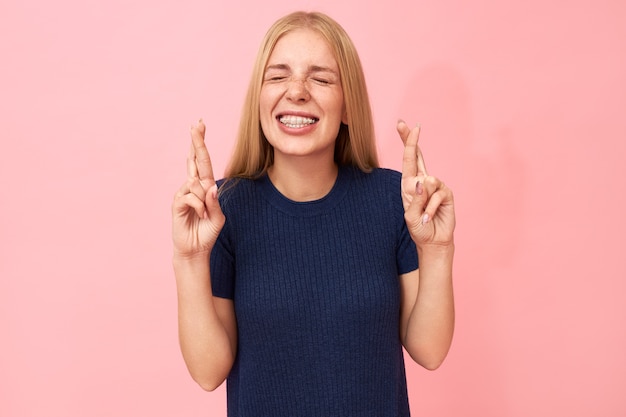 Breek een been! taille portret van grappige schattig bijgelovige tienermeisje met tanden haakjes handgebaar, kruising middelvinger over index, wensen voor geluk