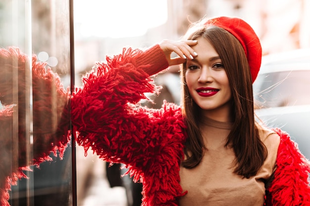 Browneyed aantrekkelijke vrouw in goed humeur poseren op straat Close-up shot van modieus meisje met rode lippenstift gekleed in lichte jas hoed en beige Tshirt