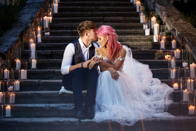 Gratis foto bruid met roze haren en stijlvolle bruidegom zitten op voetstappen met glanzende kaarsen