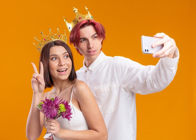 Bruidspaar bruidegom en bruid met boeket bloemen in trouwjurk dragen gouden kronen glimlachend vrolijk doen selfie met smartphone staande over oranje muur