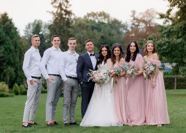 Bruidspaar en beste vrienden gekleed in modieuze bruiloft kleding staan in de rij op de groene tuin