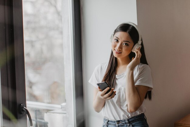 Bruinogig meisje kijkt naar de camera en houdt de telefoon vast Aziatische vrouw in witte top geniet van een liedje op een koptelefoon