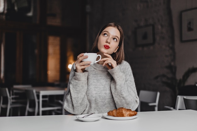 Bruinogige dame met rode lippenstift die zorgvuldig met kop thee stelt. Vrouw in grijze trui zittend aan tafel met croissant.