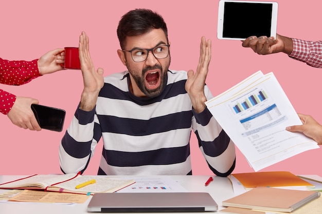 Gratis foto brunet man zit aan bureau omringd met gadgets en papieren