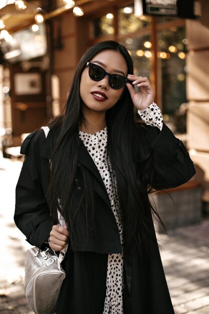 Brunette, aantrekkelijke jonge vrouw zet buiten een zonnebril op Portret van een Aziatische gebruinde dame in zwarte trenchcoat houdt een zilveren handtas