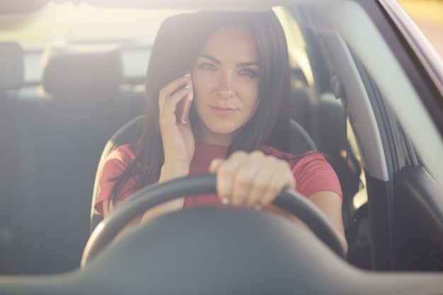 Brunette jonge vrouwelijke chauffeur spreekt via moderne mobiele telefoon tijdens het rijden auto, heeft ernstige exressie