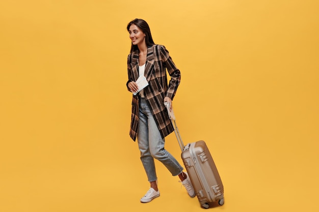 Brunette langharige vrouw in denim broek en bruine jas poses met kaartjes en koffer portret van gelukkig reizen meisje beweegt en glimlacht op geïsoleerde gele achtergrond