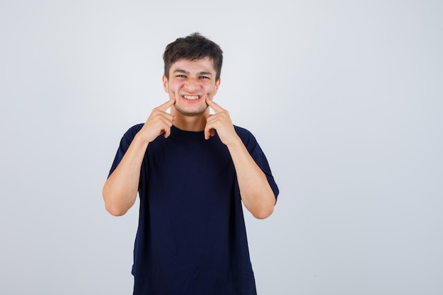 Brunette man op wangen met vingers in t-shirt te drukken en er grappig uit te zien. vooraanzicht.