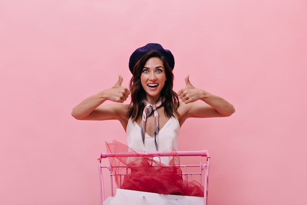 Brunette meisje in blauwe baret is blij met winkelen en duimen opdagen. Gelukkige vrouw in goed humeur in paarse hoed en witte blouse vormt op roze achtergrond.