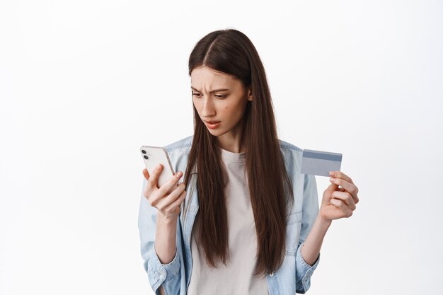 Brunette meisje kijkt verward naar het scherm van de smartphone, houdt een creditcard vast, kan niet begrijpen hoe de kaart moet worden geregistreerd om een bestelling te plaatsen, staande over een witte muur