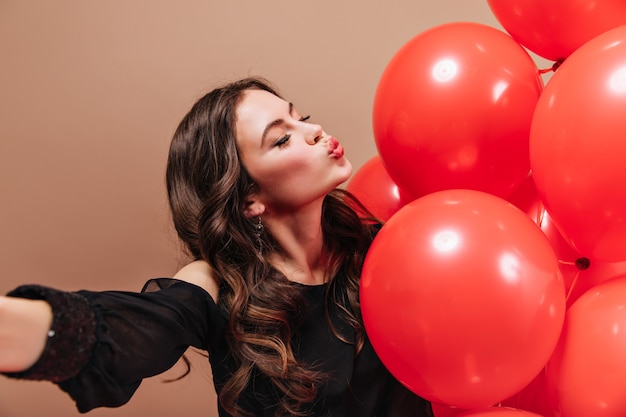 Brunette meisje met golvend haar neemt selfie, blaast kus en houdt rode ballonnen.