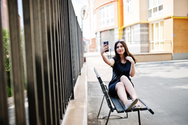Brunette meisje met zwarte jurk zonnebril zittend op een bankje luisteren naar muziek van koptelefoon telefoon en poseren in de straat van de stad