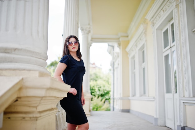 Brunette meisje op zwarte jurk zonnebril poseerde tegen oude vintage huis op straat van city