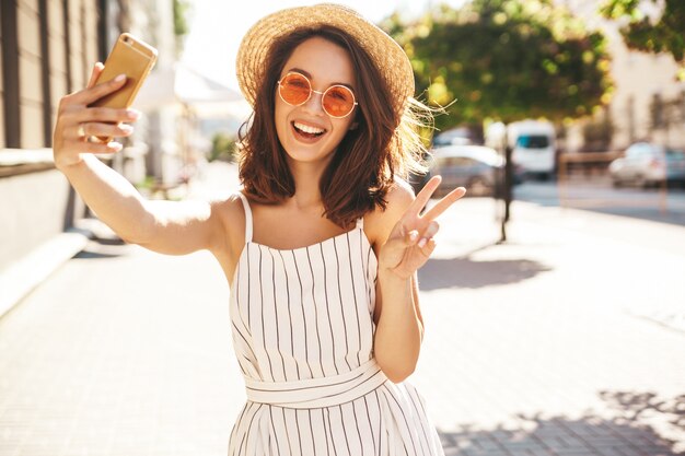 brunette model in zomer kleding poseren op straat met behulp van mobiele telefoon met vredesteken