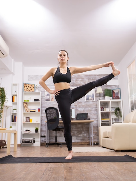 Brunette volwassen vrouw doet een staande been yoga pose in de woonkamer.