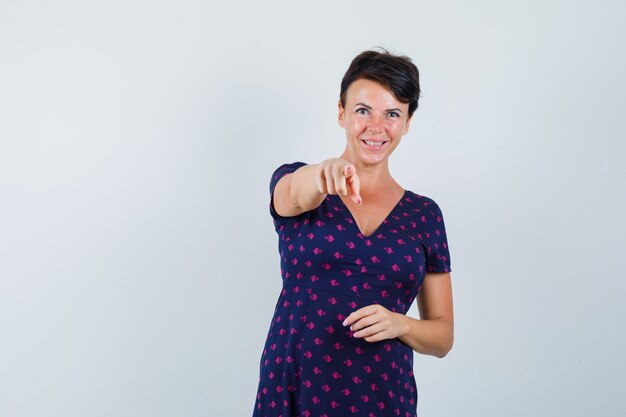 Brunette vrouw wijzend met haar wijsvingers in paars en rood patroon jurk en op zoek gelukkig, vooraanzicht.