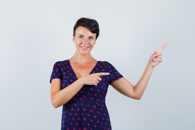 Brunette vrouw wijzend naar de rechterbovenhoek met haar wijsvingers in paars en rood patroon jurk en op zoek optimistisch, vooraanzicht.