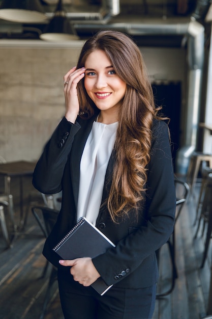 Brunette zakenvrouw met golvend lang haar en blauwe ogen staat met een notitieblok in handen en glimlacht wijd