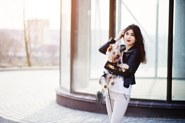 Brunette zigeunermeisje met yorkshire terrier hond poseerde tegen grote ramen huis model slijtage op leren jas en tshirt met ornament broek