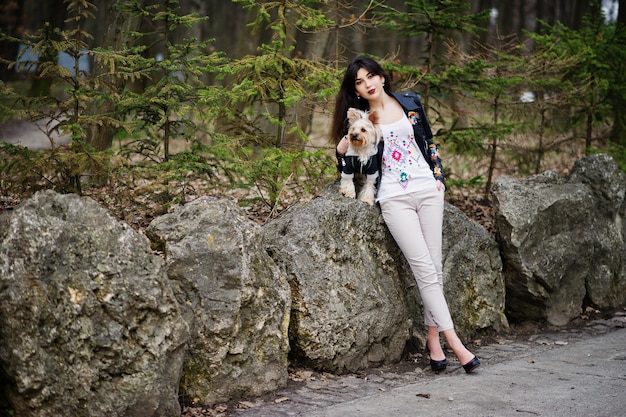 Brunette zigeunermeisje met yorkshire terrier hond poseerde tegen stenen op park Model slijtage op leren jas en tshirt met ornament broek en schoenen met hoge hakken