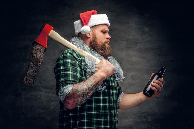 Brutaal bebaarde man in kerstmuts met groen geruit hemd, bier drinkend en houdt bijl op een schouder.