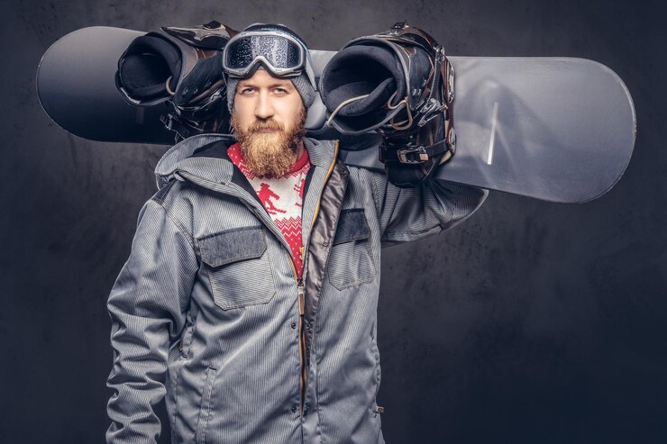 Gratis Foto brutale roodharige snowboarder met een volle baard in een wintermuts en een beschermende bril gekleed in een snowboardjas houdt een snowboard op zijn schouder in een studio. geïsoleerd op grijze achtergrond.