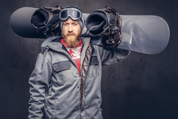 Brutale roodharige snowboarder met een volle baard in een wintermuts en een beschermende bril gekleed in een snowboardjas houdt een snowboard op zijn schouder in een studio. Geïsoleerd op grijze achtergrond.