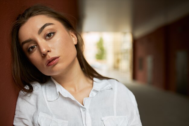 Buiten beeld van mooie stijlvolle jonge vrouw met lang losse haren poseren in boog dragen wit overhemd serieuze blik, heldere zomer straat op achtergrond.