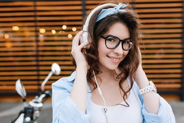 Buiten close-up portret van schattig lachend meisje in glazen witte oortelefoons aan te raken