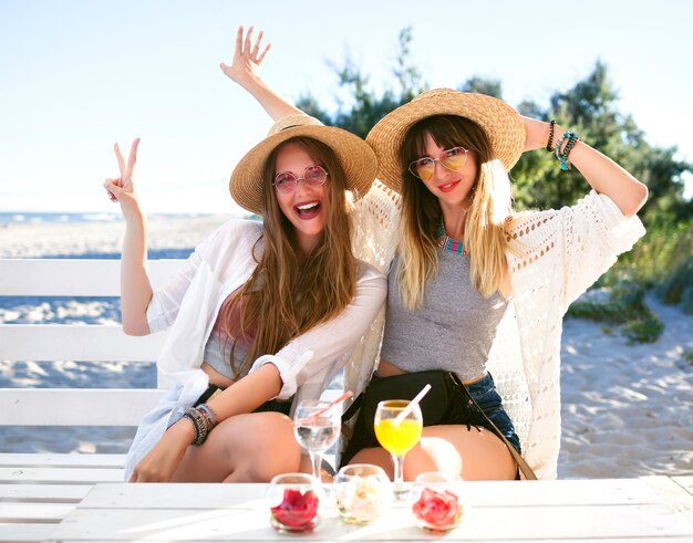 Buiten fanny portret van twee zus verslaan vrienden meisje plezier knuffels glimlachen en grimassen maken op strandbar, boho hipster kleding, lekkere cocktails drinken, oceaan zomervakantie.