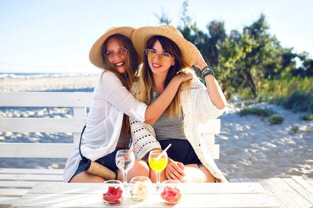 Buiten fanny portret van twee zus verslaan vrienden meisje plezier knuffels glimlachen en grimassen maken op strandbar, boho hipster kleding, lekkere cocktails drinken, oceaan zomervakantie.