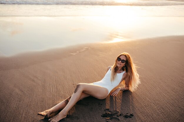 Buiten foto van glamoureuze jonge vrouw die zich voordeed op strand in hete zomerdag. Portret van mooi Kaukasisch meisje in zwarte zonnebril die op zand met geïnspireerde glimlach liggen.