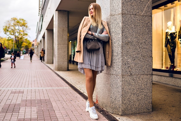 Buiten levensstijl mode portret van vrij elegante glamour blonde vrouw met lange benen, trendy sneakers, jurk trui en jas dragen, poseren in Europa stad, alleen reizen.