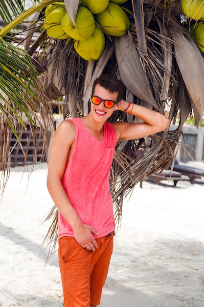 Buiten mode portret van knappe stijlvolle man geweldige tijd doorbrengen op tropisch strand, poseren in de buurt van kokospalm, heldere outfit en neon zonnebril dragen.