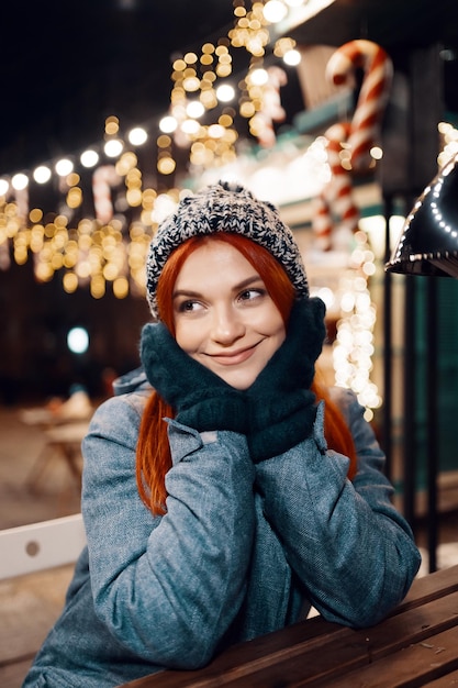 Buiten nacht foto van jong mooi gelukkig lachend meisje genieten van feestelijke decoratie, poseren in kerstmarkt, in de straat van de Europese stad, het dragen van gebreide muts