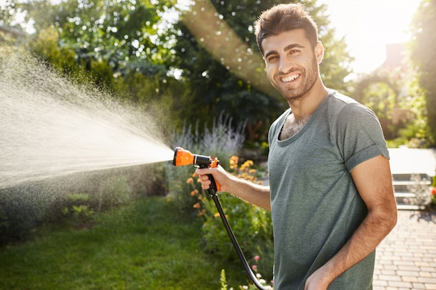 Buiten portret van jonge aantrekkelijke Kaukasische tuinman met baard en stijlvol kapsel in blauw t-shirt glimlachen, planten water geven met tuingereedschap, productieve zomerochtend.