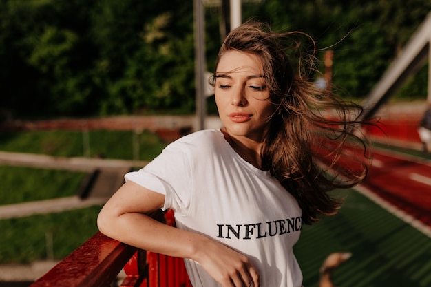 Buiten portret van stijlvolle mooie vrouw in wit t-shirt staande op de brug in zonlicht Verfijnde vrouw met vliegend haar lopen op laan met glimlach