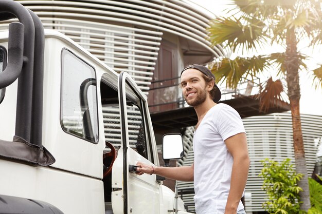 Buiten schot van jonge blanke mannelijk model met baard poseren op zijn witte crossover utility wagen, hand op handvat te houden. Stijlvolle man krijgt in zijn auto