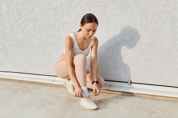 Buiten schot van jonge vrouw runner veters vastbinden, witte top en beige leggins dragen, sport oefeningen buiten doen, brunette vrouw trainen, opleiding, gezondheidszorg.