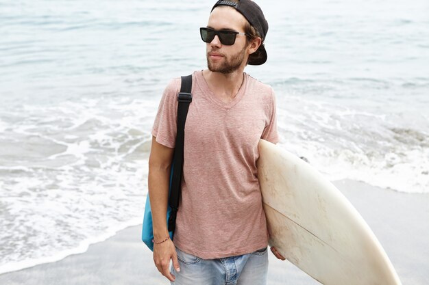 Buiten schot van modieuze jonge mannelijke model dragen zwarte tinten en snapback met witte surfplank onder zijn arm