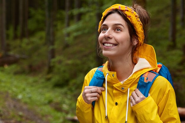 Buiten schot van vrolijke jonge vrouw kijkt bedachtzaam in de verte, draagt gele hoofdband en regenjas, dwaalt in het bos