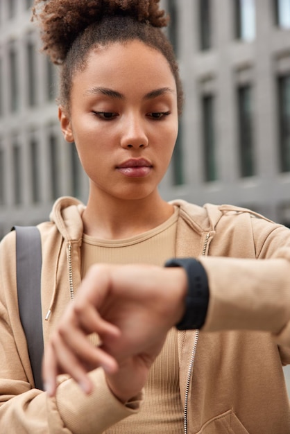 Gratis foto buiten schot van vrouw met krullend haar gekleed in sweatshirt controleert tijd op smartwatch wacht op coach oefeningen bereidt zich regelmatig voor op training poses tegen onscherpe achtergrond gezonde levensstijl