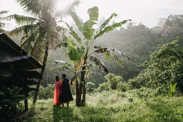 Buiten schot van vrouwelijke toerist in roze regenjas poseren met vriendje op jungle. Portret van paar koelen in tropisch woud.