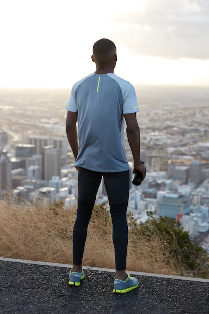 Buiten verticale shot van atletische man draagt sportkleding, staat achterover, bewondert uitzicht op de natuur en stadsgezicht van bovenaf, draagt sportfles met water, geniet van ochtendtraining. Fitness concept